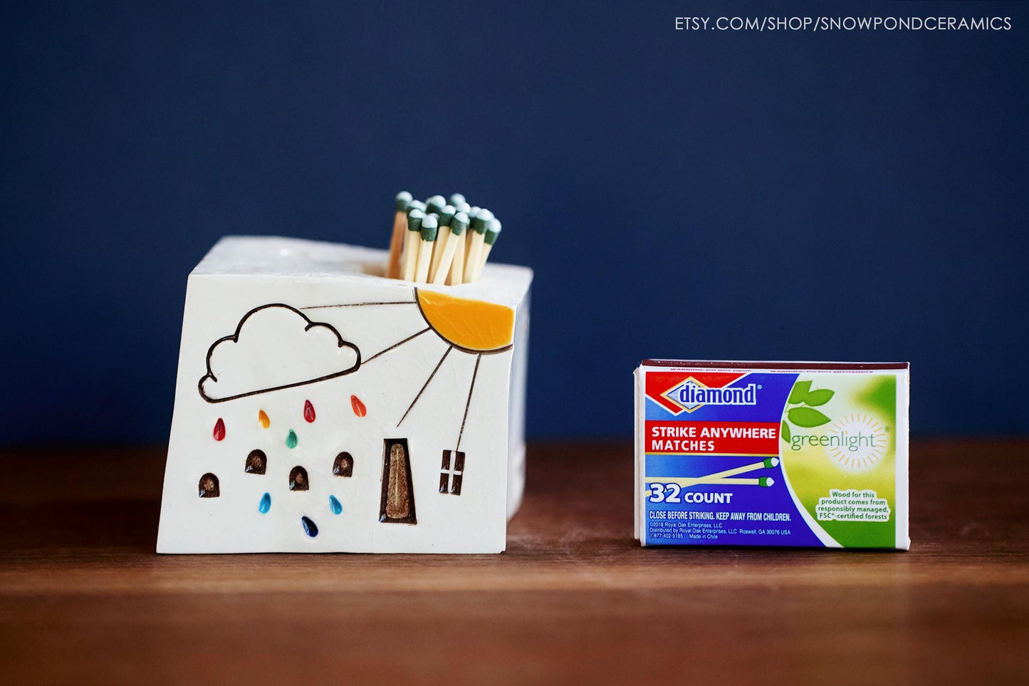 A white ceramic house shaped match striker with a cloud and rainbow raindrops on the front and a sunshine.